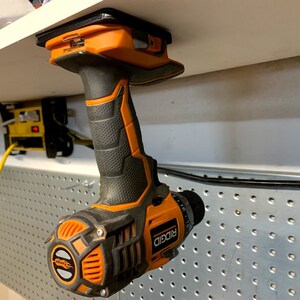 Pu&ograve; includere: Un trapano avvitatore a batteria arancione e nero con manico nero e punta in metallo argentato. Il trapano &egrave; appeso a uno scaffale bianco con un supporto magnetico.