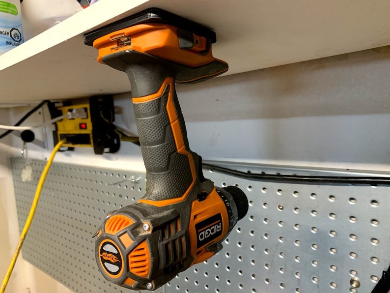 Pu&ograve; includere: Un trapano avvitatore a batteria arancione e nero con manico nero e punta in metallo argentato. Il trapano &egrave; appeso a uno scaffale bianco con un supporto magnetico.