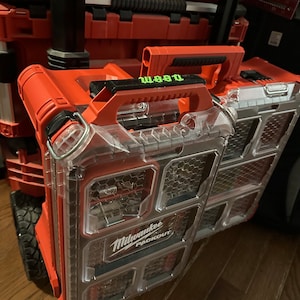 May include: A red and clear plastic Milwaukee Packout organiser with a black handle. The organiser has multiple compartments and is labelled "Milwaukee Packout".