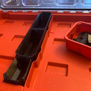May include: Black 3D-printed storage organisers inside a red toolbox. The organisers include a long, divided tray and a small square container holding additional black components. The toolbox is open, revealing its interior.
