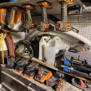 Pu&ograve; includere: Una collezione di utensili elettrici arancioni e neri, tra cui trapani, una pistola per chiodi e una sega, sono appesi a una parete con pannelli forati. Ci sono anche diverse batterie e cassette degli attrezzi su uno scaffale sottostante. Gli utensili sono tutti del marchio Ryobi.