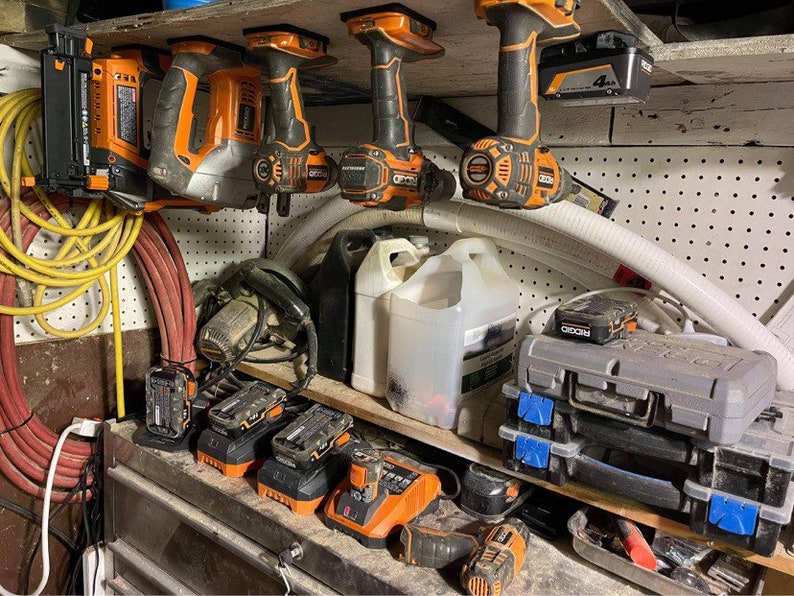 Pu&ograve; includere: Una collezione di utensili elettrici arancioni e neri, tra cui trapani, una pistola per chiodi e una sega, sono appesi a una parete con pannelli forati. Ci sono anche diverse batterie e cassette degli attrezzi su uno scaffale sottostante. Gli utensili sono tutti del marchio Ryobi.