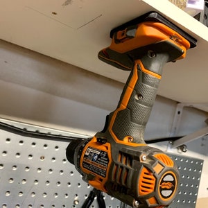 Pu&ograve; includere: Un trapano avvitatore a batteria nero e arancione con manico nero &egrave; montato su uno scaffale bianco con un supporto magnetico.