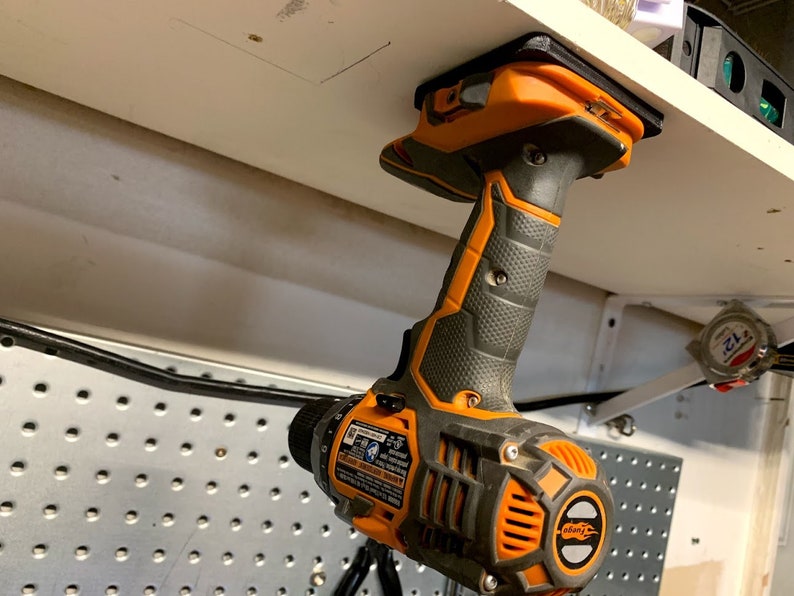 Pu&ograve; includere: Un trapano avvitatore a batteria nero e arancione con manico nero &egrave; montato su uno scaffale bianco con un supporto magnetico.