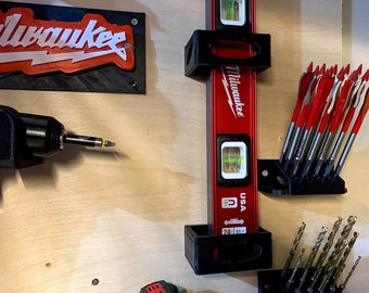 Può includere: Primo piano di un organizzatore di utensili da parete con un livello a bolla Milwaukee rosso, un trapano nero e un set di punte. L'organizzatore è in plastica nera e ha un design semplice.