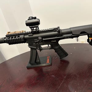 May include: Black rifle with a scope and a tactical rail, mounted on a black stand. The rifle has a pistol grip and a stock. The stand has the text "DOOM 30" in red.