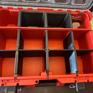 May include: Open red and black Milwaukee Packout storage container with multiple compartments. The container has a clear lid and black dividers. The Milwaukee logo is visible. A tray of gold-wrapped chocolates is in the background.