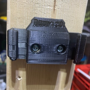 May include: A black 3D-printed wall mount with the text "ROOM3D" on top, attached to a light-colored wooden plank with screws. The mount has two slots and is designed to hold an item.