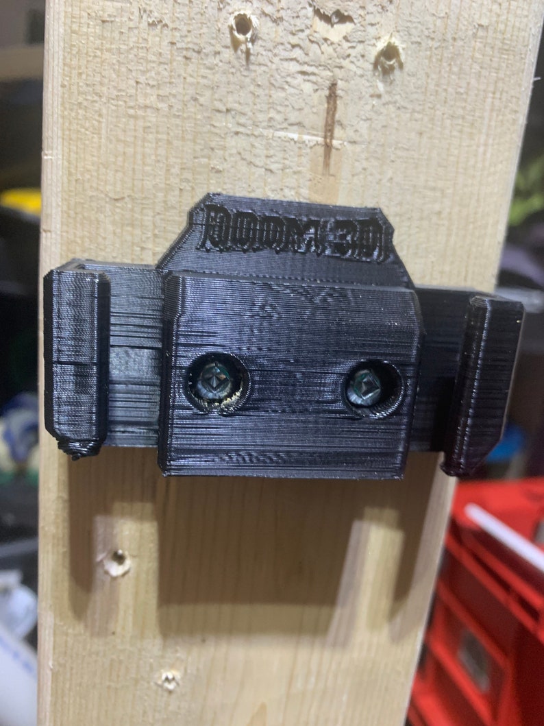 May include: A black 3D-printed wall mount with the text "ROOM3D" on top, attached to a light-colored wooden plank with screws. The mount has two slots and is designed to hold an item.