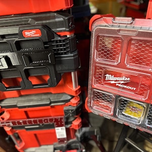 May include: A stack of red and black Milwaukee Packout toolboxes. A clear plastic organiser with red detailing and the Milwaukee Packout logo is visible. These toolboxes are designed for storage and organisation.