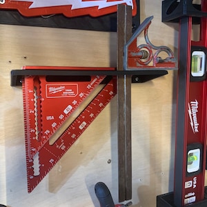 May include: A red and black Milwaukee try square with a level attached to it. The try square is mounted on a wall with a black metal bracket. The level is also mounted on the wall with a black metal bracket. The level is red and black and has a bubble level in the center. The try square has a ruler on the side with markings in inches. The level has a ruler on the side with markings in inches. The try square is labeled "Milwaukee Try Square" and the level is labeled "Milwaukee".