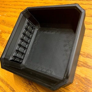 May include: A black plastic tray with a textured, grid-like pattern on one side. The tray is square shaped and has a raised edge.