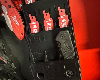 Può includere: Un organizzatore di utensili in plastica nera con sei fessure per contenere lame da sega. L'organizzatore è montato su una parete rossa. Le lame da sega sono etichettate con il testo "DIA Milwaukee" e "6/12 TPI".