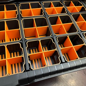 May include: A black and orange parts organizer with multiple compartments. The organizer has a grid-like structure with rectangular sections divided by orange dividers. The compartments are designed to hold and organize small items.