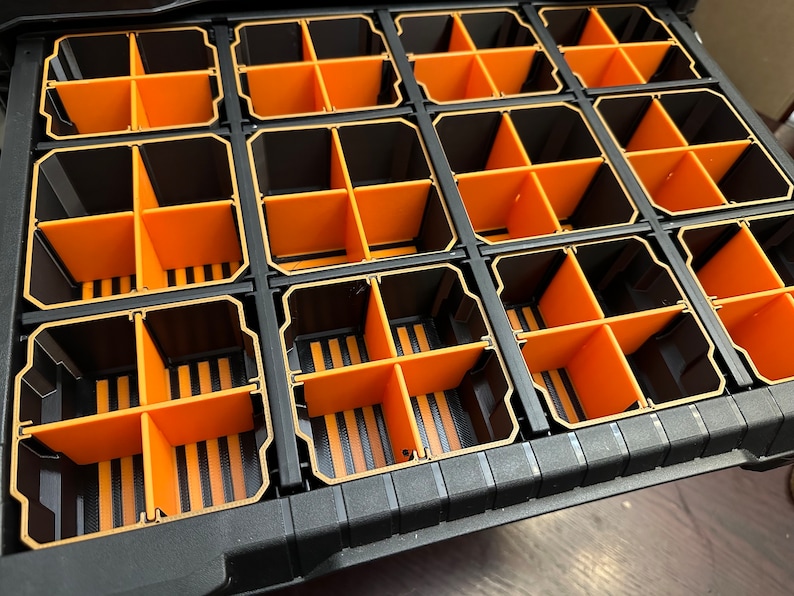 May include: A black and orange parts organizer with multiple compartments. The organizer has a grid-like structure with rectangular sections divided by orange dividers. The compartments are designed to hold and organize small items.