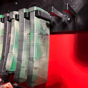 May include: Five green camouflage magazine holders with black clips attached to a black wall mount. The magazine holders have the text "BE WOOD" printed on them.