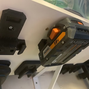 May include: A black 3D-printed power tool holder mounted on a white surface. An 18V Ridgid battery is attached to the holder. The battery is gray and black with orange accents. Other tool holders are also visible.