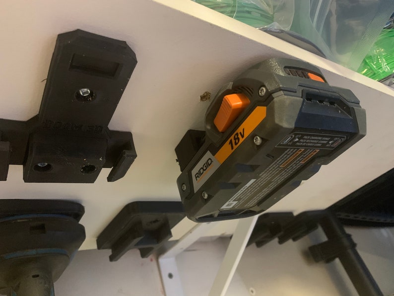 May include: A black 3D-printed power tool holder mounted on a white surface. An 18V Ridgid battery is attached to the holder. The battery is gray and black with orange accents. Other tool holders are also visible.