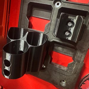 Puede incluir: Organizador de herramientas negro impreso en 3D con m&uacute;ltiples portavasos y soportes de montaje. El organizador est&aacute; fijado a una base de pl&aacute;stico roja y negra, probablemente para una caja de herramientas o un sistema de almacenamiento. El dise&ntilde;o incluye dos agujeros circulares en los portavasos.