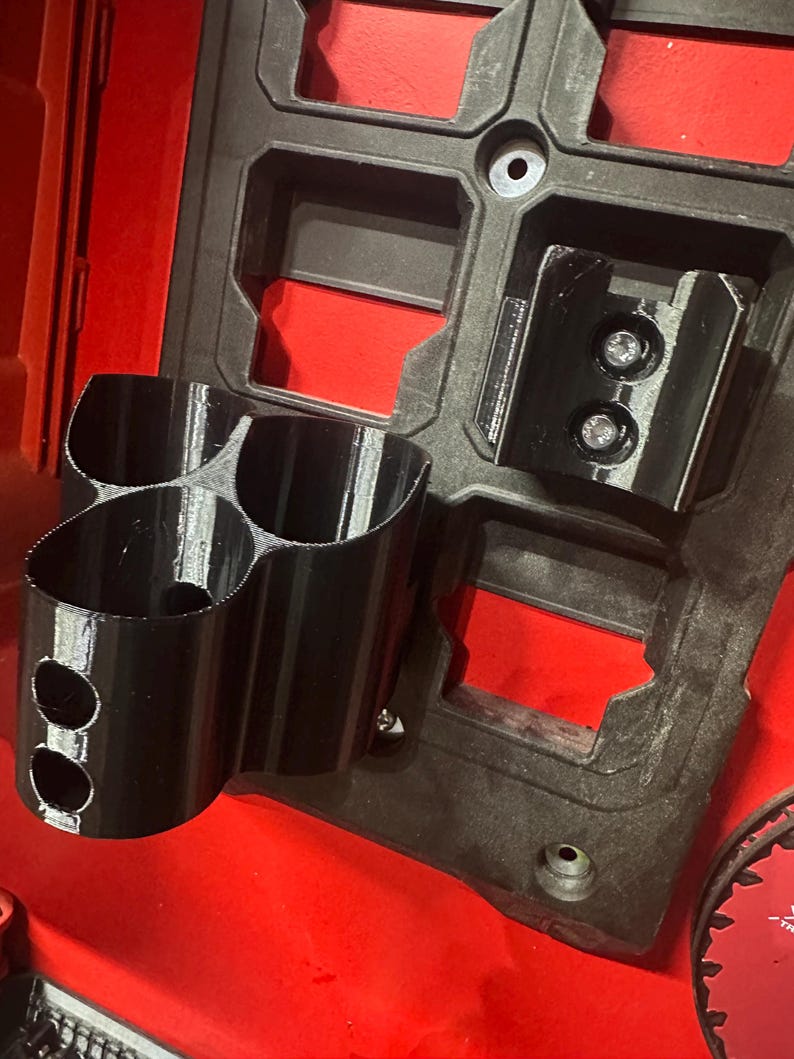 Puede incluir: Organizador de herramientas negro impreso en 3D con m&uacute;ltiples portavasos y soportes de montaje. El organizador est&aacute; fijado a una base de pl&aacute;stico roja y negra, probablemente para una caja de herramientas o un sistema de almacenamiento. El dise&ntilde;o incluye dos agujeros circulares en los portavasos.