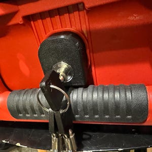 May include: Close-up of a red object with a black lock and key. The lock is mounted on the red surface, and a set of keys hangs from the lock. The object has a black, ribbed handle.
