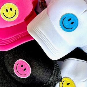 May include: Three different trucker hats with smiley face patches. One hat is pink with a yellow smiley face, one is white with a blue smiley face, and one is black with a pink smiley face.