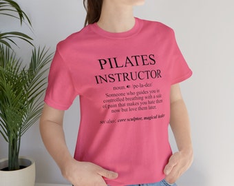 May include: Pink t-shirt with a black graphic that reads "PILATES INSTRUCTOR" and a definition of the word. The definition reads "noun. /pe-la-dez/ Someone who guides you in controlled breathing with a side of pain that makes you hate them now but love them later. see also: core sculptor, magical healer"