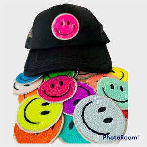May include: A black mesh trucker hat with a pink sequined smiley face patch. The hat is surrounded by a pile of colourful sequined smiley face patches.