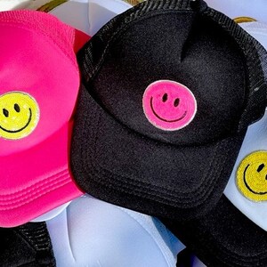May include: Three trucker hats with smiley face patches. One hat is pink, one is black, and one is white. The smiley faces are yellow, pink, and yellow.