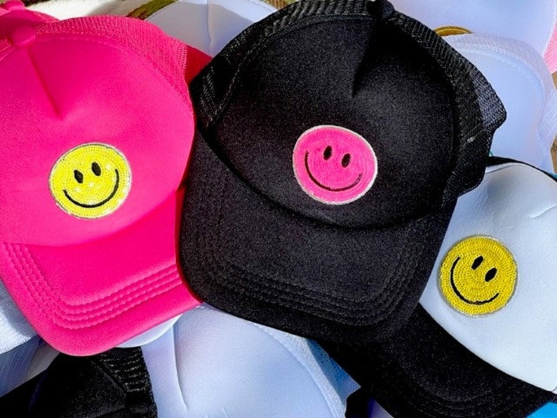 May include: Three trucker hats with smiley face patches. One hat is pink, one is black, and one is white. The smiley faces are yellow, pink, and yellow.