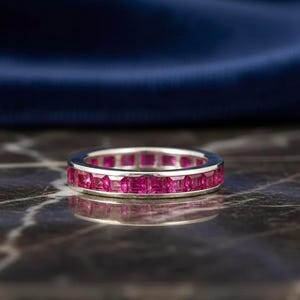 May include: A silver-toned eternity band ring featuring a continuous row of square-cut, vibrant pinkish-red gemstones set in a channel setting. The ring displays a polished metallic finish and a uniform design around the entire circumference.