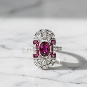 Può includere: Un anello color argento con una gemma centrale di colore rosa intenso a taglio ovale, circondata da pietre rosa rettangolari più piccole e da un bordo di pietre bianche rotonde. La montatura allungata presenta una cornice decorativa.