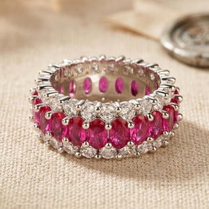 Può includere: Un anello eternity color argento con una fila centrale di pietre rosa vivaci a taglio ovale, affiancata da due file di piccole pietre bianche rotonde e trasparenti. La montatura in metallo sostiene le pietre in un motivo circolare.