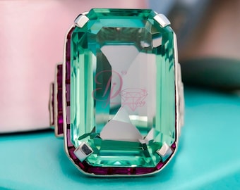 May include: A large, rectangular aquamarine-colored gemstone ring with a silver band. The ring features a large, emerald-cut center stone and smaller, rectangular ruby accents on the sides. The ring is set against a light blue background.