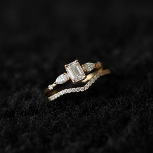 Peut inclure: Une bague de fiançailles en or avec une pierre centrale de taille émeraude, flanquée de deux diamants en forme de poire. Un anneau incurvé et délicat avec de petits diamants est en dessous. Sur fond sombre.