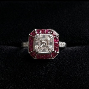 Peut inclure: Bague en argent ornée d'un grand diamant carré central, entouré d'un halo de pierres rouges. La monture est également sertie de petites pierres. L'ensemble est présenté sur un fond noir.