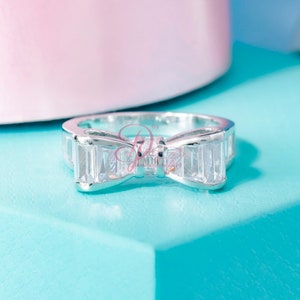 May include: A silver ring featuring a clear crystal bow design. The ring is displayed on a light blue surface.