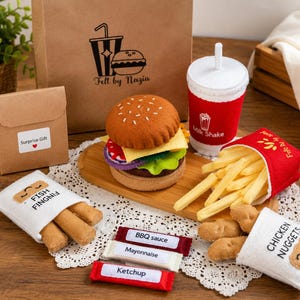 Op de afbeelding: Een set speelgoedvoedsel van vilt met een hamburger, een rood-witte milkshakebeker, een rood doosje friet, een wit doosje vissticks en een wit doosje kipnuggets. Inclusief kleine zakjes met BBQ-saus, mayonaise en ketchup.
