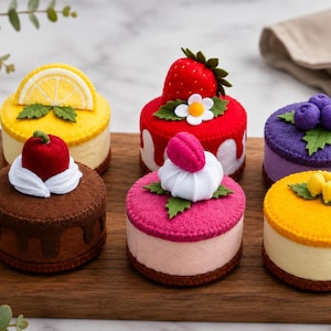 Könnte beinhalten: Sechs Miniatur-Filzkuchen auf einem Holzbrett. Die Kuchen sind mit verschiedenen Elementen verziert, darunter eine Zitronenscheibe, eine Erdbeere mit Blüte, Blaubeeren, eine Kirsche mit weißer Creme, ein rosa Macaron und gelbe Fruchtstücke.