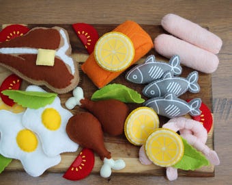 May include: A collection of felt play food items including a brown steak with a yellow butter pat, two fried eggs, two brown chicken drumsticks, an orange salmon fillet with a lemon slice, three gray fish, two pink sausages, and tomato slices.