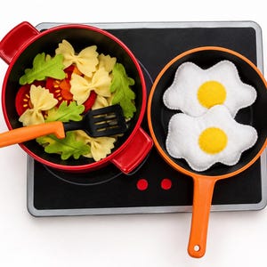 May include: A toy stovetop with a red pot containing felt bow-tie pasta, green leaves, and red tomato slices, alongside an orange pan holding two felt fried eggs. A black plastic spatula rests inside the red pot on the black and silver stovetop.