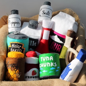 May include: A burlap sack filled with various felt food items, including a can of Heinz Baked Beans, a can of tuna chunks, a jar of honey, a jar of strawberry jam, a bottle of milk, a bottle of pepper, a bottle of salt, a bottle of olive oil, a bag of flour, and a bottle of ketchup.