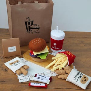 Könnte beinhalten: Ein Filz-Essensset mit einem Hamburger, Pommes Frites, Chicken Nuggets, Fischstäbchen und einem Milchshake. Das Set enthält Gewürzbeutel und eine braune Papiertüte mit dem Text "Felt by Nazia".