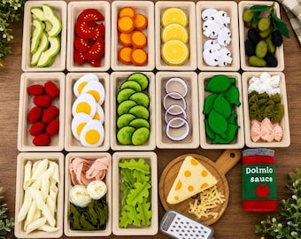 May include: A collection of felt food items including sliced avocado, tomatoes, carrots, lemons, mushrooms, olives, eggs, cucumbers, onions, spinach, pasta, lettuce, cheese on a board, and a can labeled Dolmio sauce, all in rectangular trays.