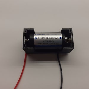 May include: A black plastic battery holder with a silver cylindrical battery inside. The battery is labelled "RCR123A 800mAh 3.7V 2.96Wh". Red and black wires are connected to the holder.
