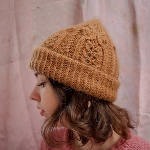 Op de afbeelding: Een vrouw draagt ​​een bruine gebreide beanie met een kabelbreipatroon. Ze draagt ​​een roze gebreide trui.