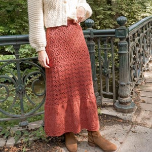 May include: A woman wearing a cream-colored, button-down cardigan and a long, rust-colored skirt with a detailed openwork pattern. She is standing near a decorative metal railing and a wooden bridge, with trees in the background.