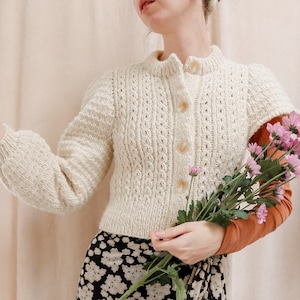 Op de afbeelding: Een crèmekleurige gebreide cardigan met een knoopsluiting en een delicaat opengewerkt patroon. De cardigan is kort en wordt gedragen met een zwart-witte bloemenrok.