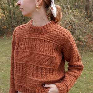 Op de afbeelding: Een roestkleurige gebreide trui met getextureerde horizontale banden. De trui heeft een ronde hals en lange mouwen. Het model draagt gouden oorbellen en een wit haaraccessoire. De achtergrond is een vage buitenscène met bomen en gras.