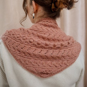 Op de afbeelding: Een close-up van een vrouw die een roze gebreide sjaal draagt met een kabelbreipatroon. De sjaal wordt gedragen over een witte trui.